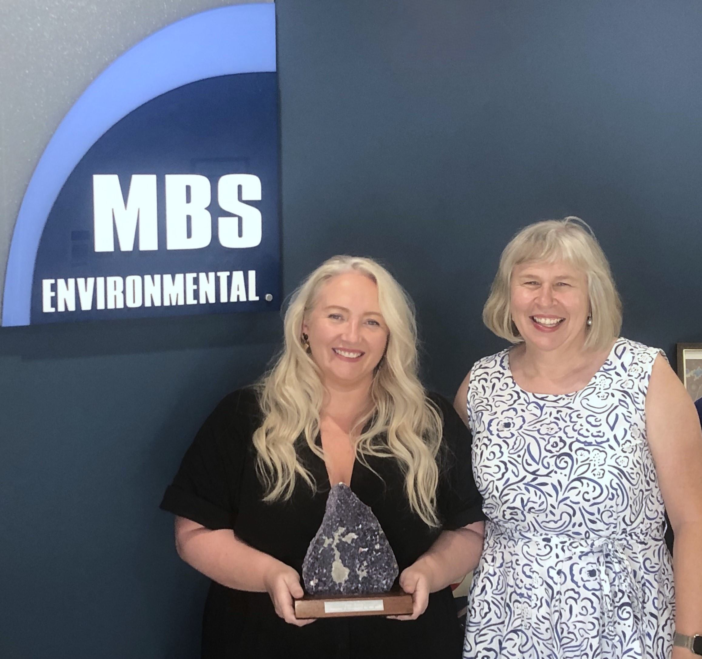 Two women: Orla Tone and Kristy Sell, standing in front of a dark background, with an MBS Environmental sign. Orla is holding an award.
